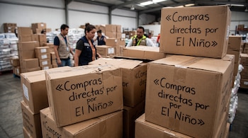 Cajas de cartón apiladas con la frase Compras directas por El Niño en un almacén con varias personas al fondo.