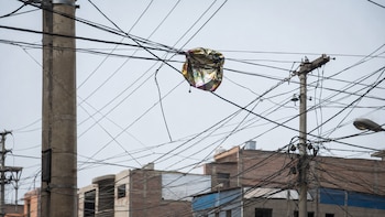 Globo metálico desinflado enredado en densa red de cables eléctricos aéreos sobre postes de concreto. Al fondo, edificios y cielo claro