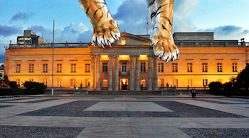 Vista frontal de la Casa de Nariño iluminada al anochecer, con dos grandes patas de tigre en acuarela sobre su techo bajo un cielo azul nublado.