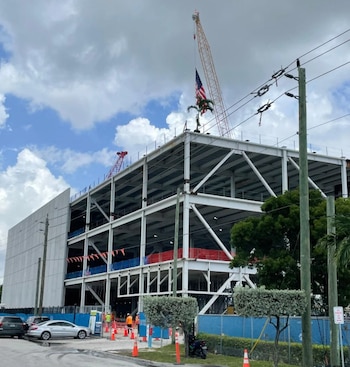 Edificio en construcción con estructura de acero, una grúa en el techo con una bandera de Estados Unidos, conos de tráfico y automóviles