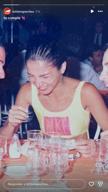 Mujer de cabello oscuro con camiseta amarilla y logo 'NIKE', riendo en una mesa con vasos y un salero, junto al perfil de un hombre. Fondo borroso