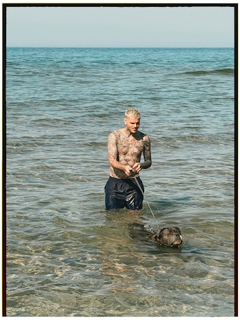 Un hombre caucásico musculoso y tatuado con cabello rubio blanqueado está de pie en el mar hasta la cintura, sosteniendo una correa de perro. Un perro oscuro nada frente a él