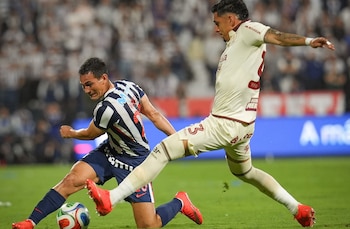 Un futbolista con uniforme azul y blanco disputando el balón frente a otro jugador con uniforme crema en una cancha de césped