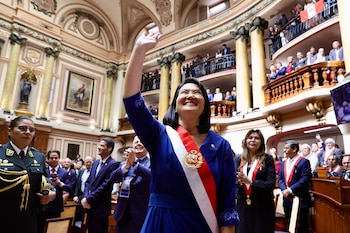 Una mujer sonriente con traje azul y banda presidencial roja y blanca levanta la mano. Está en un gran salón con columnas, balcones y cuadros, rodeada de personas