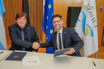 Dos hombres de negocios se estrechan las manos sobre una mesa con documentos. Al fondo, banderas de Argentina, la Unión Europea y CONICET