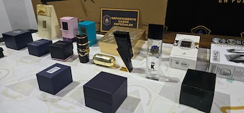 Varios perfumes y cajas de productos falsificados, como fragancias de lujo y una cortadora de pelo Wahl, sobre una mesa. Fondo con cartel policial