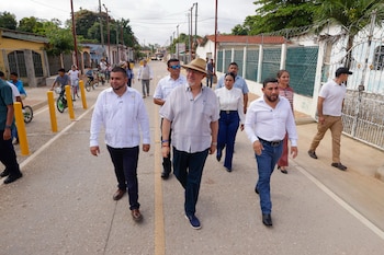 Un grupo de personas camina por el centro de una calle pavimentada con viviendas a los lados y varios ciclistas al fondo
