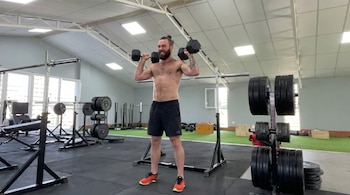 Un hombre con barba y moño, sin camiseta, en shorts negros, realiza press de hombros con mancuernas de pie en un gimnasio con equipo de pesas.