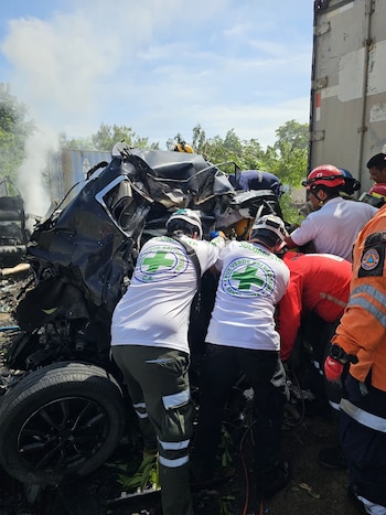 Varios socorristas con uniformes blancos y rojos inspeccionan un automóvil negro destruido junto a la estructura de un camión