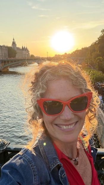 Primer plano de mujer con cabello rizado y gafas de sol rojas, sonriendo. De fondo se ven un río, un puente y edificios al atardecer con el sol
