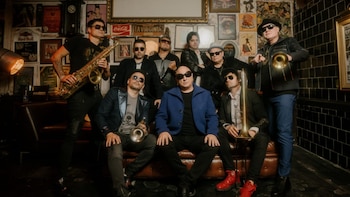 Siete hombres, integrantes de la banda La Mosca Tsé-Tsé, posan en una habitación con paredes decoradas. Un músico tiene un saxofón, otros dos sostienen trompetas