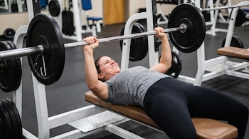 Mujer de 30 años acostada en un banco, sosteniendo una barra con discos de peso arriba de su pecho en un gimnasio con equipo deportivo.