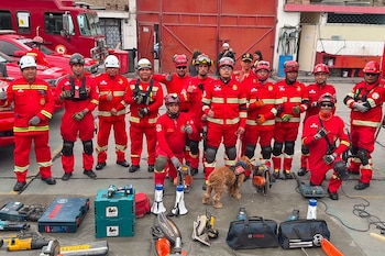 Un grupo de bomberos en uniformes rojos y cascos posa con herramientas de rescate y un perro en primer plano, frente a camiones de bomberos