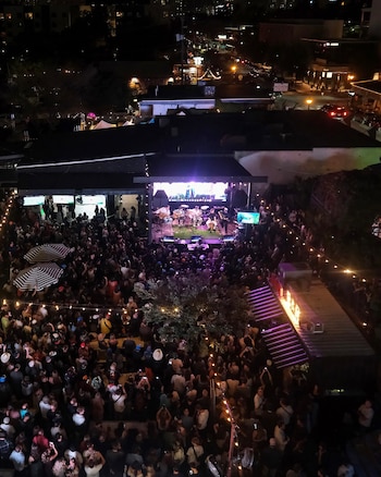 Vista en picado de una banda sobre un escenario iluminado ante una multitud en un patio exterior con guirnaldas de luces por la noche