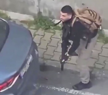 Hombre con corte de pelo corto, camisa oscura y pantalón claro, sosteniendo un rifle y llevando una mochila marrón, parado junto a la parte trasera de un coche