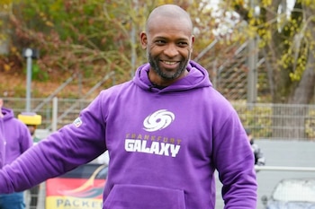 Hombre sonriente con cabeza rapada y barba, viste una sudadera morada con el logotipo de Frankfurt Galaxy. Fondo borroso con árboles y una valla