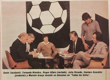 Un grupo de hombres sentados y de pie alrededor de una mesa en un estudio con la ilustración de un balón de fútbol al fondo