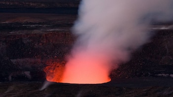 Vista aérea de un cráter volcánico oscuro, con un intenso resplandor naranja-rojo en su interior y una densa columna de humo blanco elevándose