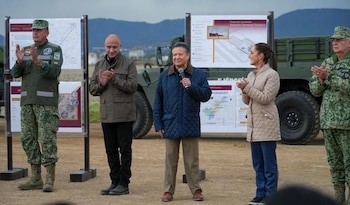 Cinco personas, incluyendo militares y civiles, de pie al aire libre frente a paneles informativos. Un hombre habla por un micrófono mientras otros aplauden