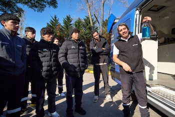 Siete hombres de pie al aire libre. Un hombre sostiene una botella con líquido azul. Una furgoneta azul abierta está detrás. Hay árboles y cielo azul