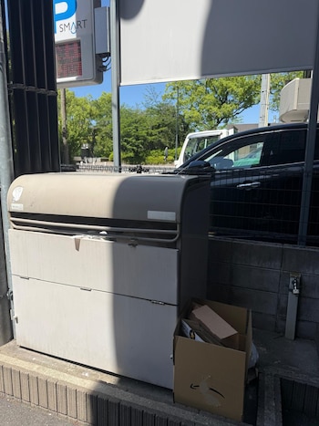 Primer plano de una caja de servicios urbana gris al lado de una caja de cartón con objetos reciclables en un bordillo. Al fondo, un coche negro