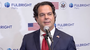 Un hombre con traje azul marino y corbata roja habla frente a un micrófono ante un panel blanco con el logotipo de Fulbright Perú