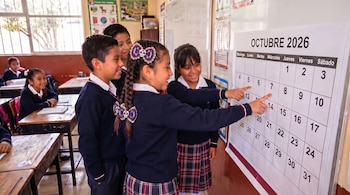 Cuatro estudiantes vistiendo uniforme azul y a cuadros señalan un calendario impreso de octubre de 2026 pegado en la pared.
