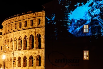 Fachada de piedra con arcos de un anfiteatro romano iluminado y pared de un edificio con proyecciones azules y dos ventanas encendidas