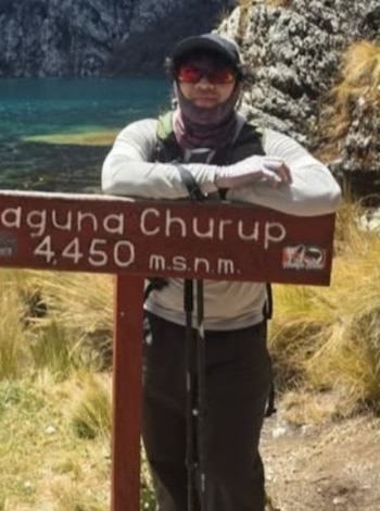 Un hombre con gorra, gafas de sol y ropa de montaña se apoya en un letrero de madera que dice Laguna Churup con una laguna de fondo