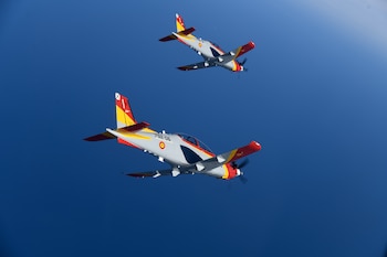 Dos aviones de instrucción grises, rojos y amarillos con hélices en vuelo, con insignias de España, contra un cielo azul claro