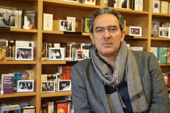 Primer plano de Juan Gabriel Vásquez, un hombre de mediana edad con gafas y bufanda gris, delante de estanterías de madera llenas de libros