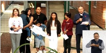 Un grupo de seis adultos y tres niños de pie al aire libre; uno de los niños sostiene una llave gigante de papel; un hombre habla por micrófono; recuadro con intérprete de señas
