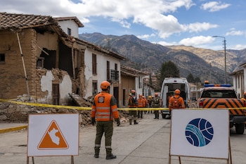 Personal de rescate con chalecos naranjas y cascos junto a una casa de adobe derrumbada, vehículos de emergencia y carteles institucionales.