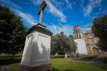 Una cruz de piedra sobre un pedestal blanco, un campanario con cúpula roja, la fachada de una iglesia, árboles y césped bajo cielo azul con nubes