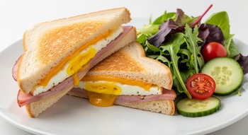 Sándwich caliente de jamón, queso y huevo, cortado en dos, con queso derretido y yema de huevo visible, acompañado de ensalada verde con pepino y tomate cherry.