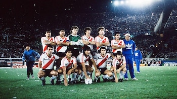 Once futbolistas con uniforme blanco y franja roja posan de pie y agachados junto a un balón en una cancha de césped con público en las tribunas