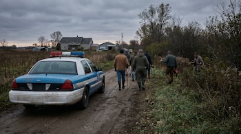 Patrullero policial blanco y azul en camino de tierra rural. Un grupo de personas camina por la derecha, buscando. Fondo con vegetación densa, campo y una finca, bajo cielo nublado.