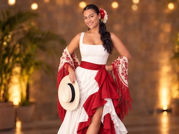 Mujer con vestido blanco y rojo, mantón bordado y flores en el cabello, que sostiene un sombrero de paja frente a un fondo iluminado