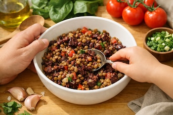 Dos manos, una de adulto y otra de niño, sujetan un bol blanco con lentejas y sangrecita, usando una cuchara. Sobre una mesada de madera, hay tomates, espinacas, ajo y aceite.