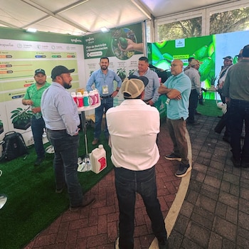 Grupo de hombres en un stand de exposición con banners de Syngenta. Se observan botellas de productos agrícolas sobre una mesa, pasto artificial y baldosas