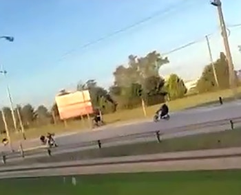 Personas a bordo de motocicletas transitan por una calle asfaltada junto a postes de luz, árboles y un cartel publicitario