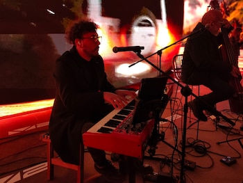 Un hombre con gafas toca un piano eléctrico rojo frente a un micrófono en un escenario con luces rojas y pantallas al fondo