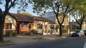 Vista de una calle residencial con dos casas de techos de teja y paredes claras, ambas con carteles de 'ALQUILER', árboles y un coche aparcado.