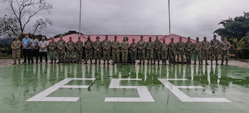 Vista frontal de un grupo de militares y civiles de pie sobre un helipuerto verde con letras blancas 'ESE', frente a un edificio con techo rojo y selva al fondo