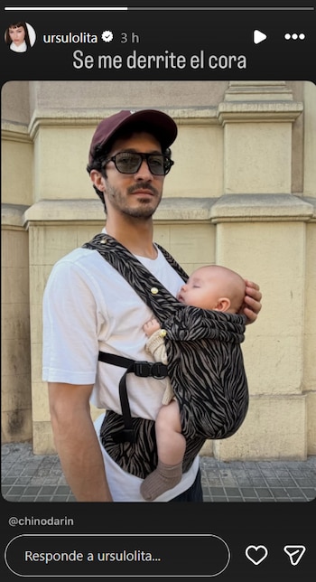 Hombre con gorra, gafas de sol y barba lleva un bebé dormido en un portabebés con estampado blanco y negro frente a una pared de edificio