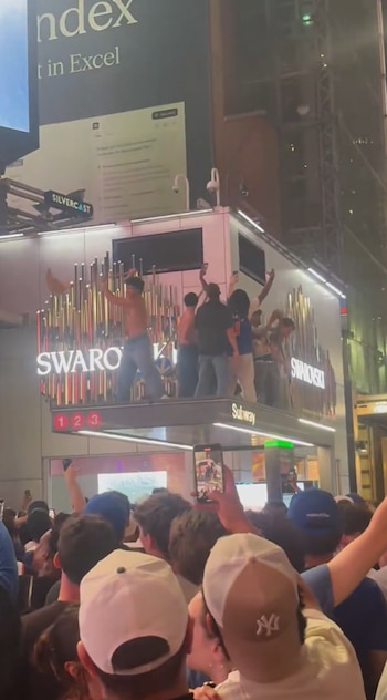 Multitud celebra en las calles de Nueva York por la noche, algunos subidos a la marquesina de una estación de metro y la tienda Swarovski iluminada