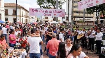 Vista de una multitud celebrando el Día de la Madre en Cúcuta, con puestos de mercado, músicos, edificios coloniales y una pancarta rosada.