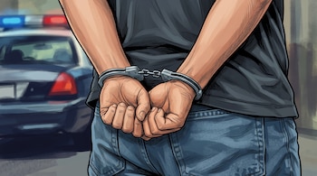 Una persona con las manos esposadas a la espalda y un automóvil policial con luces de emergencia en el fondo.
