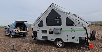 Una caravana plegable blanca con paneles solares, una camioneta azul abierta y una antena satelital en un terreno pedregoso