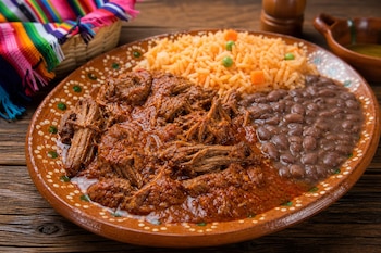 Un plato de barro servido con carne deshebrada en salsa roja, arroz y frijoles enteros sobre una mesa de madera.